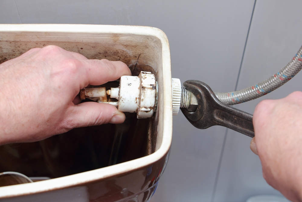 The plumber's hands unscrew the filling pipe nut from old dirty water fill valve in the empty drain toilet tank