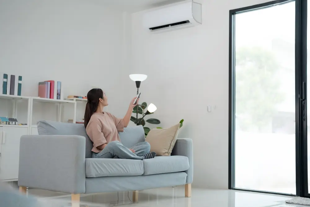 Young woman relaxes on a couch, stretching out her arms with a remote in hand, as she enjoys the cool air from an air conditioning unit in a bright, modern living room