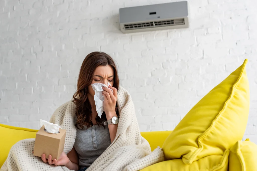 sick woman with paper napkin warming under blanket while sitting under ductless air conditioner at home